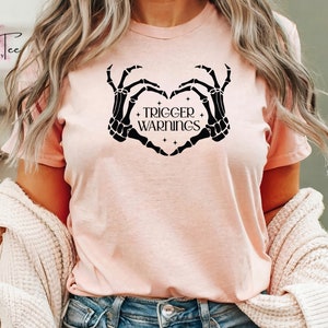 May include: A pink t-shirt with a black skeleton hand design forming a heart shape. The text "Trigger Warnings" is inside the heart.