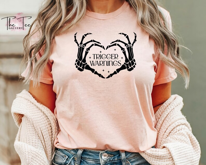 May include: A pink t-shirt with a black skeleton hand design forming a heart shape. The text "Trigger Warnings" is inside the heart.