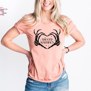 May include: A peach-colored t-shirt with a black skeleton hand design making a heart shape. The text "TRIGGER WARNINGS" is inside the heart.