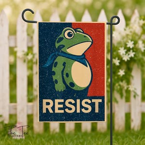 May include: A garden flag featuring a cartoon frog with a blue scarf, set against a red and blue background. The word "RESIST" is printed in bold letters. The flag is displayed on a black metal stand in a garden setting.