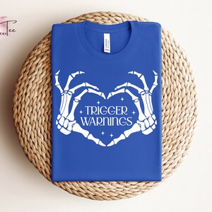 May include: A royal blue t-shirt with a white skeleton hand design making a heart shape. The text "TRIGGER WARNINGS" is inside the heart.