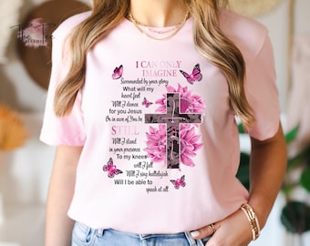 May include: Light pink t-shirt featuring a floral cross design with the text "I Can Only Imagine." The design includes pink flowers and butterflies. The shirt is a short-sleeved crew neck.