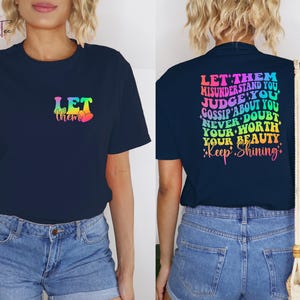 May include: A navy blue t-shirt with a rainbow gradient design that says "Let them" on the front and "Let them misunderstand you, judge you, gossip about you, never doubt your worth, your beauty. Keep shining." on the back.