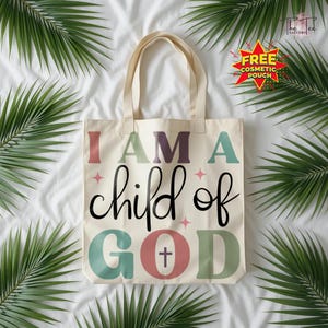 May include: Cream-colored tote bag with the text "I AM A child of GOD" in various colors. The bag has a simple design with a cross symbol in the word "GOD". The bag has two handles and is surrounded by green palm leaves.