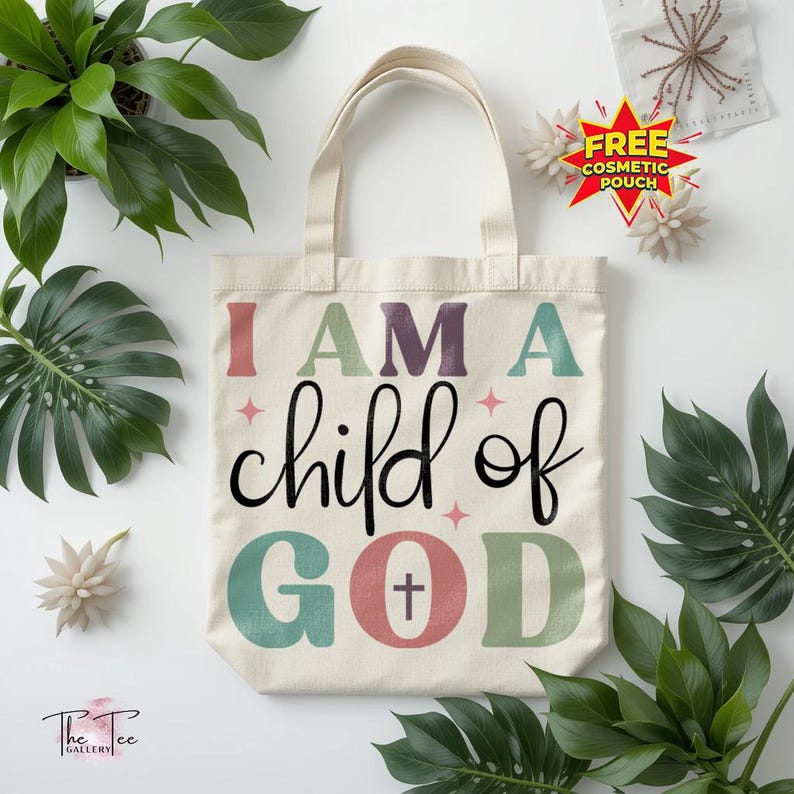 May include: Cream-colored tote bag with the text "I AM A child of GOD" in various colors and fonts. The bag features a cross symbol within the word "GOD". The bag has two handles and is surrounded by green plants and white flowers.