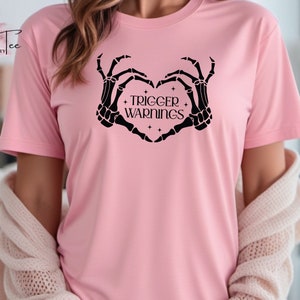May include: Pink t-shirt with a black skeleton hand design making a heart shape with the text "TRIGGER WARNINGS" inside the heart.