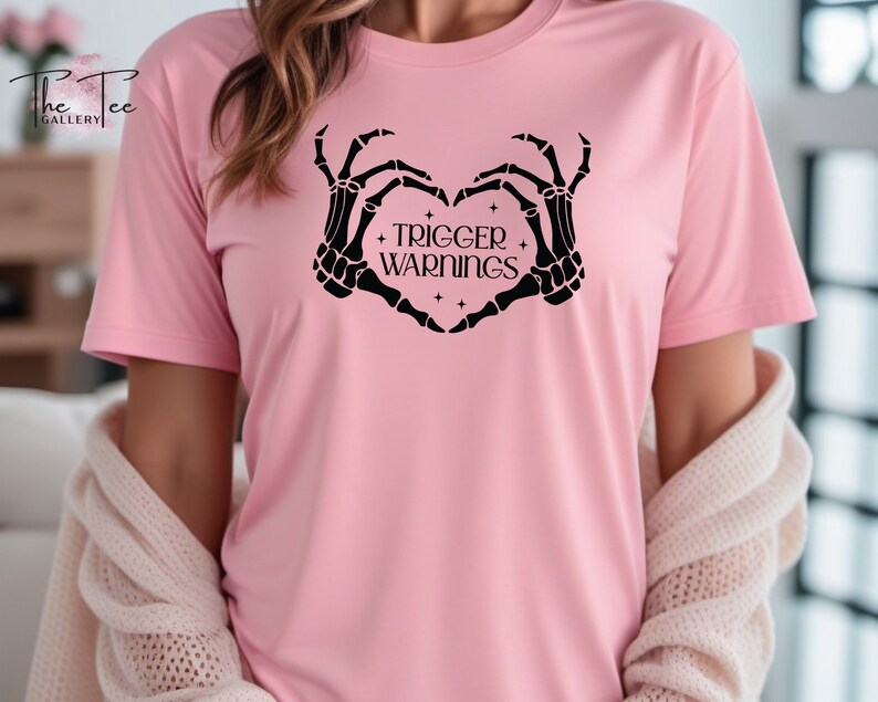 May include: Pink t-shirt with a black skeleton hand design making a heart shape with the text "TRIGGER WARNINGS" inside the heart.