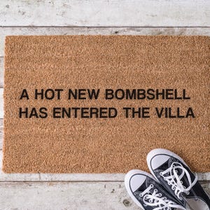 May include: A rectangular doormat made of natural coir fiber, with the phrase "A HOT NEW BOMBSHELL HAS ENTERED THE VILLA" in black lettering. A pair of black and white canvas sneakers rests on the wooden surface.