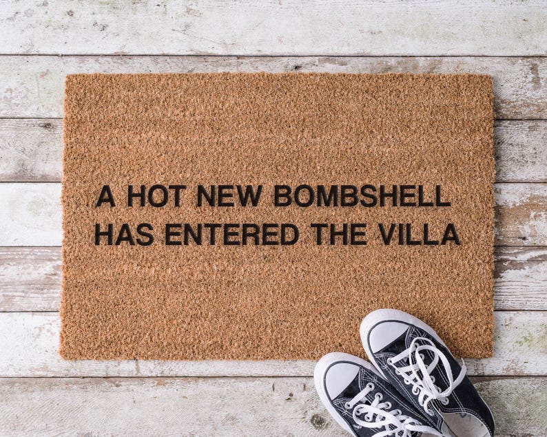 May include: A rectangular doormat made of natural coir fiber, with the phrase "A HOT NEW BOMBSHELL HAS ENTERED THE VILLA" in black lettering. A pair of black and white canvas sneakers rests on the wooden surface.