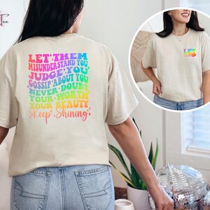 May include: A beige t-shirt with a rainbow-colored graphic on the back that says "Let them misunderstand you, judge you, gossip about you, never doubt your worth, your beauty, Keep Shining."