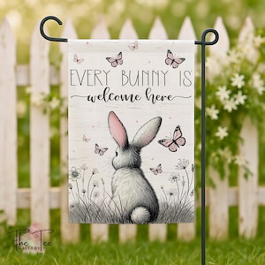 May include: A garden flag with a grey bunny and pink-lined ears, surrounded by butterflies and wildflowers. The flag has the text "Every Bunny Is Welcome Here". The flag is displayed on a black metal stand in a garden setting.