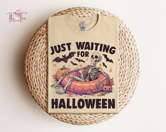 May include: A cream-coloured t-shirt featuring a graphic of a skeleton lounging in a pink inflatable pool ring. The text reads "JUST WAITING FOR HALLOWEEN" in dark brown letters. The design includes orange pumpkins, black bats, and small stars.