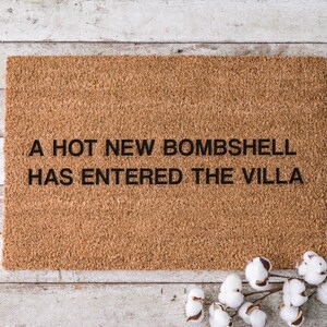 May include: A rectangular doormat made of natural coir fiber. The mat features the phrase "A HOT NEW BOMBSHELL HAS ENTERED THE VILLA" in black block letters. A sprig of cotton adds a decorative touch.