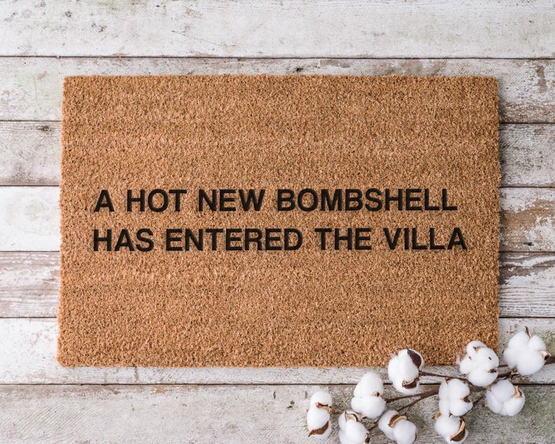 May include: A rectangular doormat made of natural coir fiber. The mat features the phrase "A HOT NEW BOMBSHELL HAS ENTERED THE VILLA" in black block letters. A sprig of cotton adds a decorative touch.