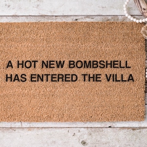 May include: A rectangular doormat with the text "A HOT NEW BOMBSHELL HAS ENTERED THE VILLA" in black letters. The mat is a light brown color and has a textured surface. The background is a light wood.