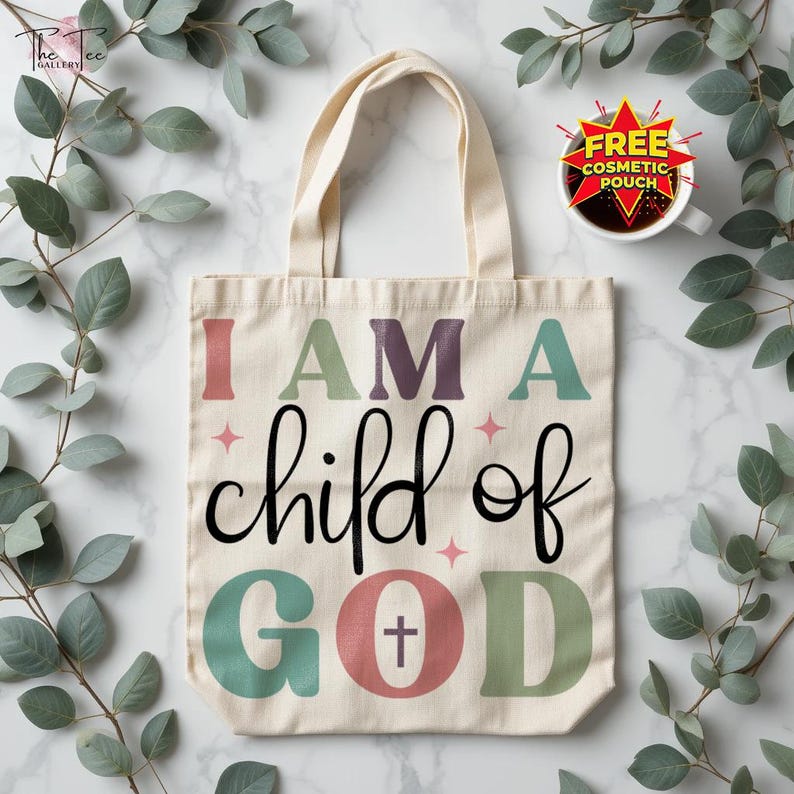 May include: Cream-colored tote bag with the text "I AM A child of GOD" in various colors. The design includes a cross within the word "GOD". The bag has two handles and is surrounded by green leaves.