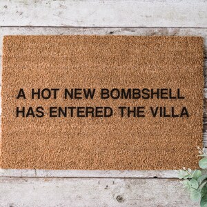 May include: A rectangular brown coir doormat with the text "A HOT NEW BOMBSHELL HAS ENTERED THE VILLA" in black letters. The doormat is placed on a light-colored wooden surface.