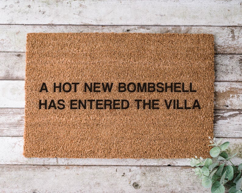 May include: A rectangular brown coir doormat with the text "A HOT NEW BOMBSHELL HAS ENTERED THE VILLA" in black letters. The doormat is placed on a light-colored wooden surface.