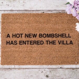 May include: A rectangular doormat made of brown coir fiber. The text "A HOT NEW BOMBSHELL HAS ENTERED THE VILLA" is printed in black. The mat is placed on a weathered, light-colored wooden surface, with a bouquet of flowers in the upper right corner.
