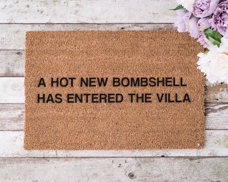 May include: A rectangular doormat made of brown coir fiber. The text "A HOT NEW BOMBSHELL HAS ENTERED THE VILLA" is printed in black. The mat is placed on a weathered, light-colored wooden surface, with a bouquet of flowers in the upper right corner.