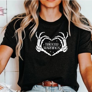 May include: Black t-shirt with a white skeleton hand design making a heart shape with the text "TRIGGER WARNINGS" inside the heart.
