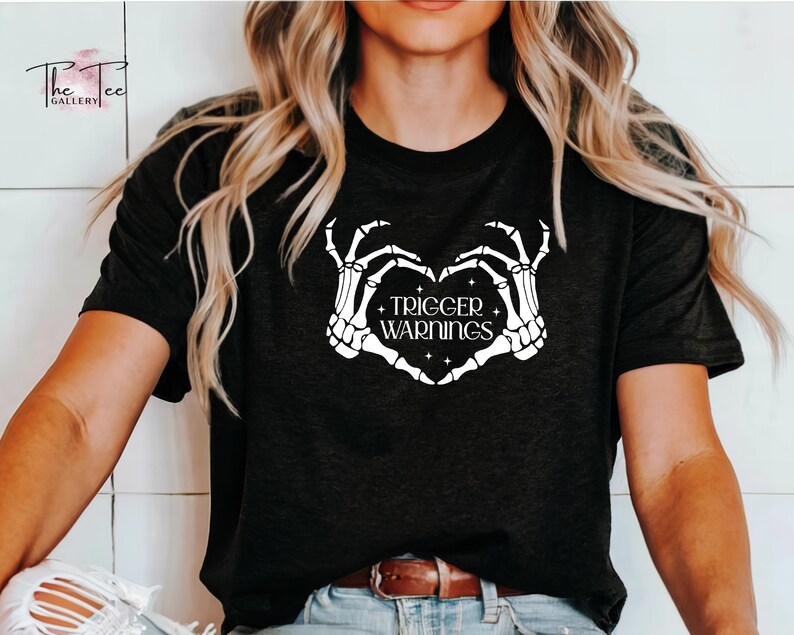 May include: Black t-shirt with a white skeleton hand design making a heart shape with the text "TRIGGER WARNINGS" inside the heart.