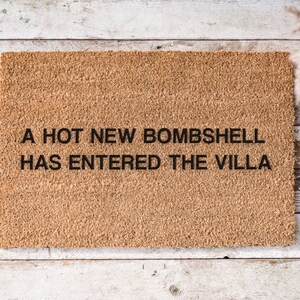 May include: A rectangular doormat made of natural coir fiber. The mat features the phrase "A HOT NEW BOMBSHELL HAS ENTERED THE VILLA" in black, centered on the mat. The doormat is placed on a light-colored wooden surface.