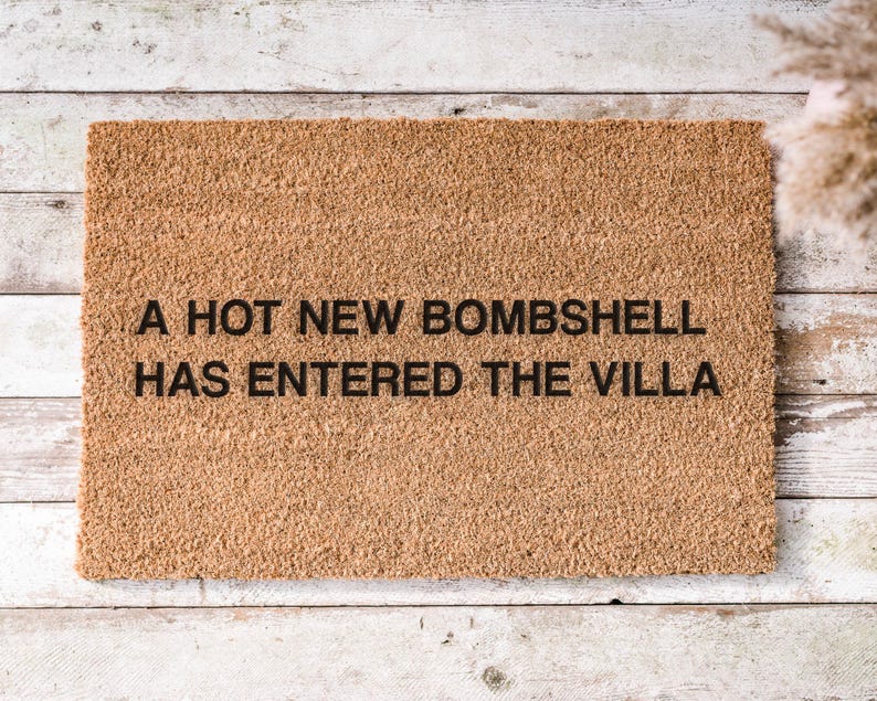 May include: A rectangular doormat made of natural coir fiber. The mat features the phrase "A HOT NEW BOMBSHELL HAS ENTERED THE VILLA" in black, centered on the mat. The doormat is placed on a light-colored wooden surface.
