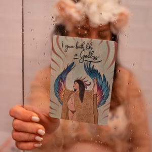 May include: A card with the text "I give birth like a Goddess" and an illustration of a woman with angel wings. The card is held in front of a person. The illustration features a woman with brown hair and a patterned shawl.