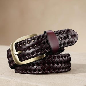 May include: A dark brown braided leather belt featuring a matte gold-toned metal buckle and a smooth leather keeper loop. The belt is coiled, showcasing the intricate woven pattern of the leather straps and the rectangular shape of the buckle.