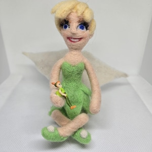 May include: A handmade felt doll of Tinkerbell, with blonde hair, blue eyes, and a green dress. The doll is holding a miniature Peter Pan figure. The doll has white wings and green shoes with white polka dots. The background is white.