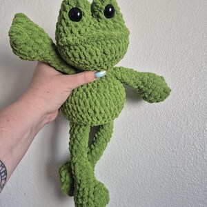 May include: A handmade green crochet frog plush toy with large black eyes. The frog has outstretched arms and long legs, giving it a playful appearance. The plush is made of a soft, textured yarn.