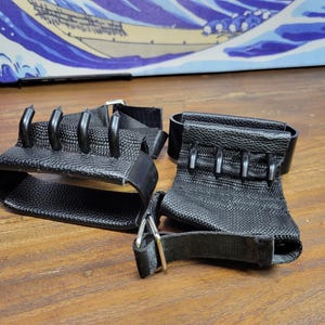 May include: Black weightlifting grips with metal hooks. The grips have a textured fabric base with adjustable straps and metal buckles. The hooks are evenly spaced and curved. The grips are designed to enhance grip strength during workouts.