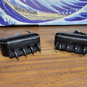 May include: A pair of black leather wrist cuffs with four claw-like spikes. The cuffs have a textured leather exterior and a buckle closure. The background features a wooden surface and a Japanese wave art print.