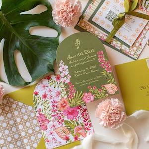 May include: Wedding stationery set featuring a Save the Date card with floral illustrations and text. The design includes pink flowers, green leaves, and a gold script. Additional cards and envelopes are also visible.
