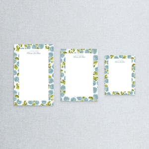 May include: Three different sizes of notepads with a blue and green floral border. The notepads have the text "A Note From Olivia De Bias" printed at the top.