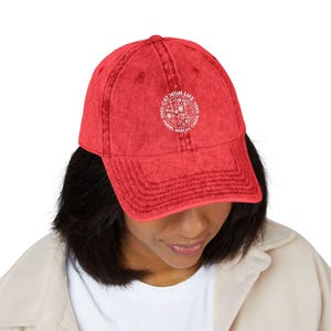 May include: Red baseball cap with a curved peak. The cap has white text and a circular graphic that reads "Cat Mom Life" and "Purrs, Snacks, Chaos". The cap is worn by a person.