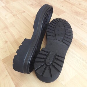 K&ouml;nnte beinhalten: Zwei schwarze Schuhsohlen mit einem robusten Profilmuster werden auf einer hellen Holzoberfl&auml;che gezeigt. Eine Sohle ist auf der Seite positioniert, wodurch das Innendesign sichtbar wird, w&auml;hrend die andere flach liegt.