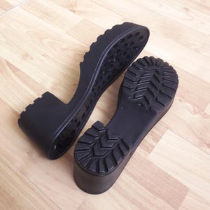 May include: Two black platform shoe soles with a textured tread pattern. The soles are made of a durable material and feature a thick heel and a ridged design for added grip. The soles are designed for footwear.