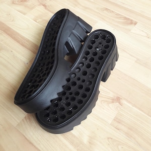 May include: Two black shoe soles with a chunky, ridged design. The soles have a series of circular cutouts and are made of a durable material. The soles are designed for a platform shoe.