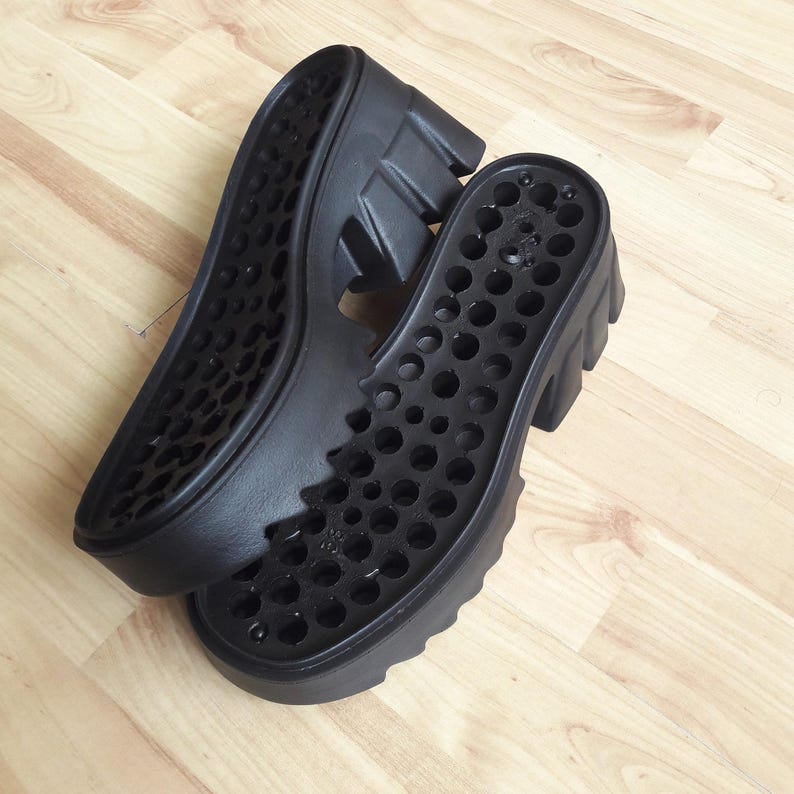 May include: Two black shoe soles with a chunky, ridged design. The soles have a series of circular cutouts and are made of a durable material. The soles are designed for a platform shoe.