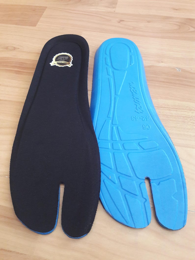 May include: Two tabi-style insoles, one black and one blue, are displayed on a wooden surface. The black insole has a logo and a blue trim. The blue insole has a textured design and text that reads "12-13".