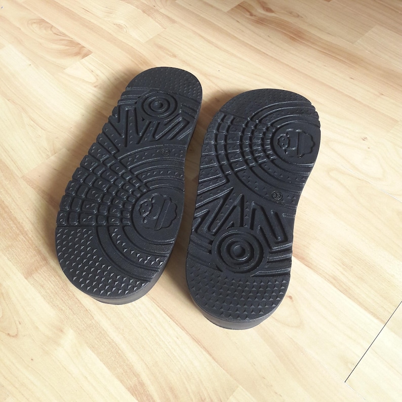 May include: Two black rubber soles of sandals, showcasing intricate tread patterns. The soles are designed with textured surfaces, including geometric shapes and a central logo. The sandals are placed on a light-colored wooden floor.