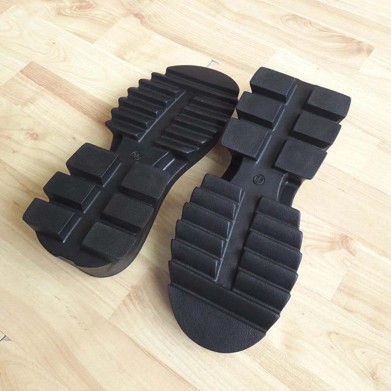 May include: Two black rubber shoe soles with a chunky, ridged tread pattern. The soles are designed for boots or heavy-duty footwear, offering grip and durability. The soles are lying on a light-coloured wooden surface.