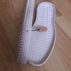 Op de afbeelding: Een paar witte rubberen schoenzool, een met een getextureerd profiel en de andere met een glad oppervlak. De zolen liggen op een houten oppervlak.