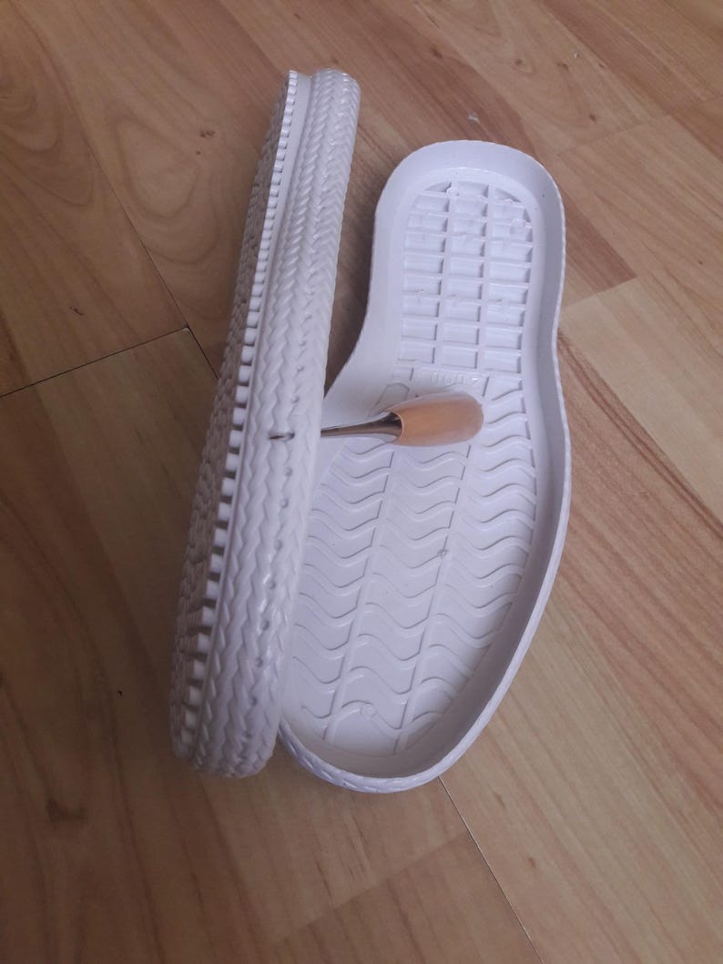 Op de afbeelding: Een paar witte rubberen schoenzool, een met een getextureerd profiel en de andere met een glad oppervlak. De zolen liggen op een houten oppervlak.