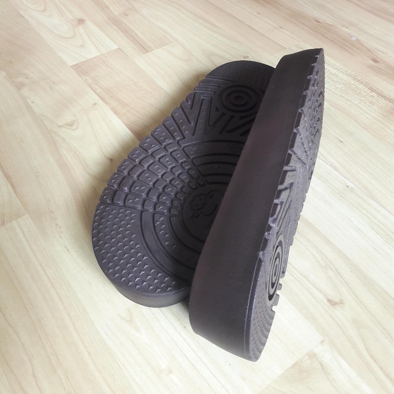 May include: Two dark brown shoe insoles with textured soles are displayed on a light wood surface. The insoles are thick and have a contoured shape, designed for comfort and support. The soles feature various patterns for grip.
