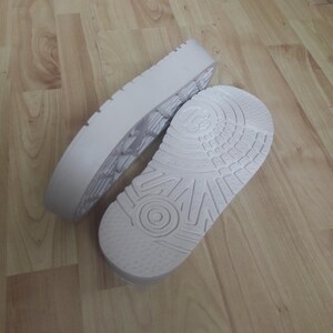 May include: Two white shoe soles, one with a textured tread and the other with a detailed pattern, are displayed. The soles are made of a light-colored material and are positioned on a wooden surface, suggesting a focus on footwear components.