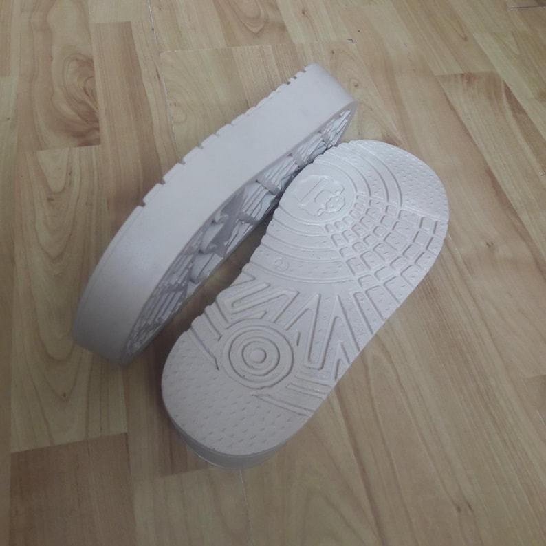May include: Two white shoe soles, one with a textured tread and the other with a detailed pattern, are displayed. The soles are made of a light-colored material and are positioned on a wooden surface, suggesting a focus on footwear components.