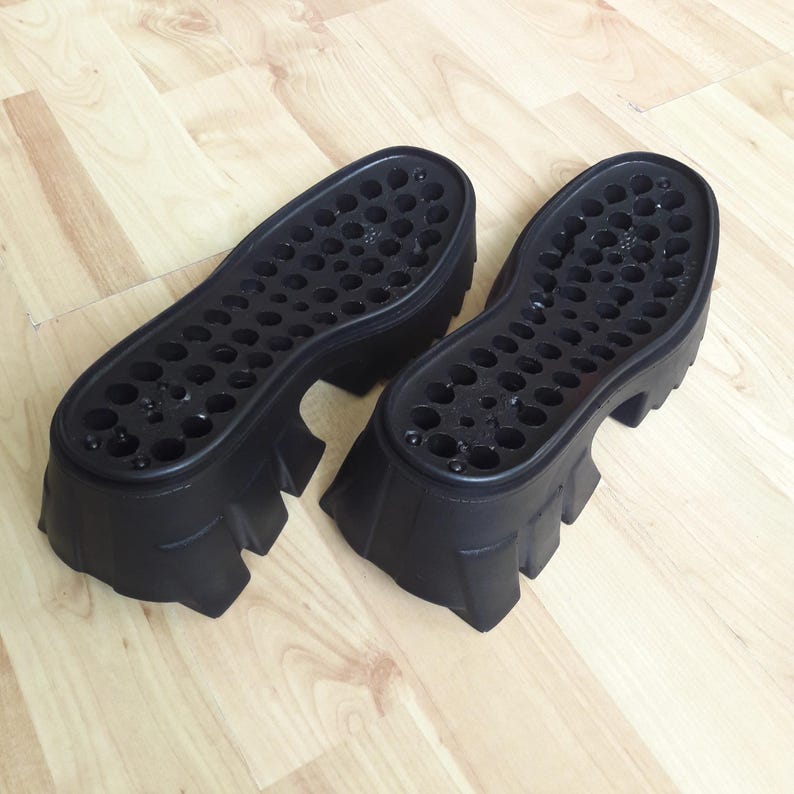 May include: Two black platform shoe soles. The soles have a chunky design with a series of small, circular holes across the surface. The soles are made of a durable material. The soles are approximately 10 cm high.